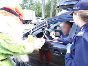 umundurowana policjantka wręcza kierowcy samochodu ulotkę. Obok niej stoi osoba przebrana za skrzata z koszem wypełnionym cukierkami