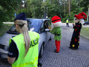 na pierwszym planie znajduje się umundurowana policjantka w kamizelce odblaskowej. Policjantka stoi przed znajdującym się na parkingu samochodem osobowym. Osoba przebrana za skrzata trzyma kosz z cukierkami i częstuje nimi kierowcę. Za nią stoi osoba przebrana za czarownicę.