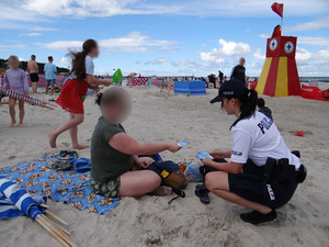 na plaży w pobliżu stanowiska ratowników wodnych policjantka w białej koszulce polo z napisem policja i czarnych spodenkach wręcza plażowiczce ulotkę