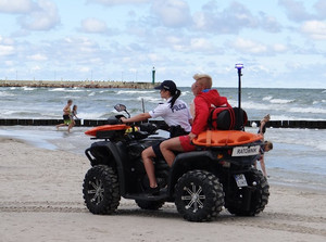 na plaży wzdłuż linii brzegowej quadem jedzie policjantka. Za nią na miejscu pasażera siedzi ratownik wodny