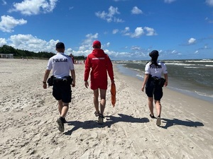 plażą wzdłuż linii brzegowej idzie patrol składający się z dwojga policjantów i ratownika wodnego