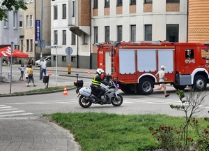 ulicą jedzie policjant na motocyklu. Przed nim biegnie zawodnik. Mijają wóz straży pożarnej