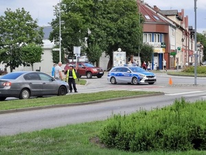 na rondzie znajduje się oznakowany radiowóz, przed rondem na drodze stoi policjantka ruchu drogowego, która rozmawia z dwoma mężczyznami