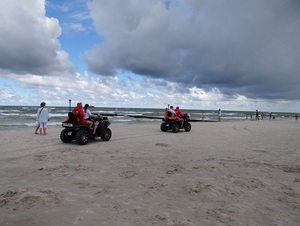 plażą wzdłuż brzegu jadą dwa quady. Jeden jest prowadzony przez policjantkę, za którą siedzi ratownik WOPR, drugi prowadzi ratownik, za nim siedzi policjant