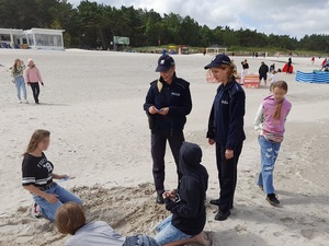 na plaży dwie uśmiechnięte policjantki rozmawiają z dziećmi bawiącymi się piaskiem