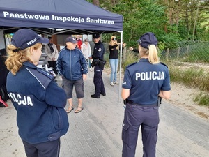 przed wejściem na plażę przy stoisku sanepidu młoda kobieta stoi z założonymi alkogoglami. Obok stoją policjanci oraz pracownicy sanepidu