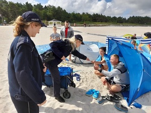 na plaży przed namiotem pod którym siedzą plażowicze stoją dwie policjantki. Jedna z nich wręcza ulotkę uśmiechniętej kobiecie