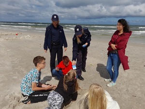 na plaży stoją dwie policjantki. Jedna z nich wyciąga rękę, w której trzyma ulotkę w stronę dzieci bawiących się na piasku