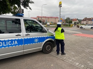 na chodniku obok oznakowanego radiowozu stoi policjantka ruchu drogowego, która obserwuje ruch pojazdów w okolicy przejścia dla pieszych