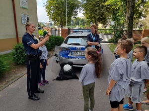 na zewnątrz przed szkołą dwie policjantki rozmawiają z grupą dzieci. Jedna stoi przy radiowozie, druga prezentuje dzieciom kajdanki