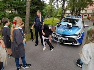 przed oznakowanym radiowozem dziewczynka ubiera policyjny sprzęt ochrony indywidualnej. Obok niej stoi uśmiechnięta policjantka