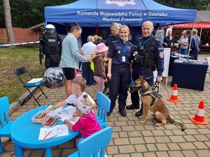 przed stoiskiem profilaktycznym KWP w Gdańsku stoją policjantka, policjant i pies służbowy. Przed nimi przy stoliku siedzą dzieci, które wykonują prace plastyczne