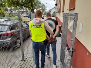 policjant w kamizelce z napisem policja wyprowadza z budynku komendy mężczyznę skutego kajdankami. Kierują się w stronę nieoznakowanego radiowozu