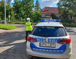 w rejonie szkoły, nieopodal przejścia dla pieszych stoi policjantka ruchu drogowego. Obok niej zaparkowany jest oznakowany radiowóz