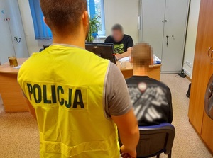 na krześle przed biurkiem siedzi zatrzymany mężczyzna. Za nim stoi policjant w kamizelce odblaskowej z napisem policja. Przy biurku siedzi policjant, który przesłuchuje zatrzymanego