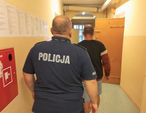 korytarz pomieszczenia dla osób zatrzymanych. W kierunku otwartych drzwi celi idzie mężczyzna, za nim idzie policjant służby dyżurnej