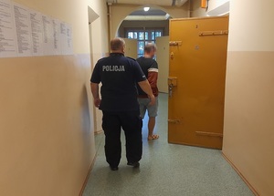 korytarz pomieszczenia dla osób zatrzymanych. Do celi wchodzi mężczyzna. Za nim znajduje się policjant służby dyżurnej