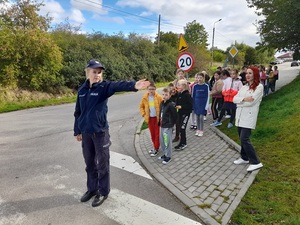 przed przejściem dla pieszych stoi grupa uczniów. Przed nimi znajduje się policjantka z wyciągniętą w bok lewą ręką