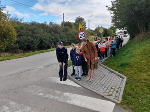 przed przejściem dla pieszych stoi grupa uczniów. Jest z nimi policjantka, która lekko pochylona do przodu tłumaczy dzieciom w pierwszym rzędzie zasady poruszania się po przejściu dla pieszych