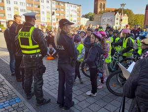 na placu stoją policjant ruchu drogowego i policjantka prewencji którzy rozmawiają ze zgromadzonymi dziećmi - uczestnikami rajdu