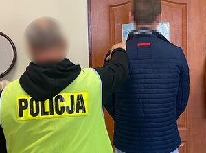 przy drzwiach stoi zatrzymany mężczyzna. Za nim znajduje się policjant w odblaskowej kamizelce z napisem policja