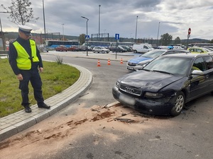 policjant ruchu drogowego przygląda się uszkodzeniom czarnego osobowego audi. Samochód ma wgnieciony przód. Droga jest posypana substancją neutralizującą wycieki płynów eksploatacyjnych