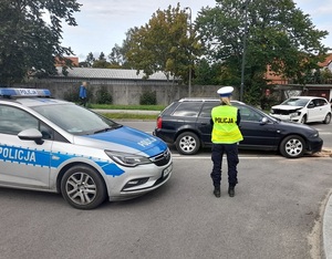 na drodze pomiędzy oznakowanym radiowozem, i czarnym audi stoi policjantka ruchu drogowego. Przed nią znajduje się biały vw z uszkodzonym przodem