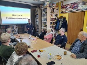 w bibliotece przy stole siedzą uczestnicy debaty, w tym umundurowany policjant. Naprzeciw przy ekranie, na którym wyświetla się napis &quot;krótki film o rzeczach oczywistych&quot;, stoi przemawiająca do zebranych policjantka