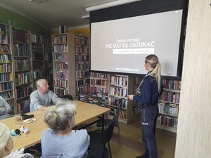 w bibliotece przy stole siedzą uczestnicy debaty. Naprzeciw przy ekranie, na którym wyświetla się napis Policja ostrzega nie daj się oszukać metodą na wnuczka, stoi przemawiająca do zebranych policjantka