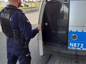 przy otwartych tylnych drzwiach oznakowanego radiowozu stoi umundurowany policjant, który przytrzymuje drzwi. W środku, za kratami siedzi zatrzymana osoba