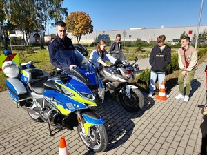 na motocyklu siedzi młoda uśmiechnięta dziewczyna. Po jej prawej stronie przy drugim motocyklu stoi policjant, po jej lewej stronie stoją trzej uśmiechnięci młodzi mężczyźni