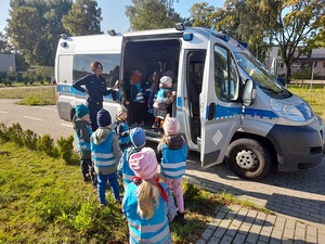 przy otwartych drzwiach radiowozu typu bus stoi uśmiechnięta policjantka. Do radiowozu wchodzą przedszkolaki