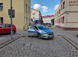 radiowóz patrolu ruchu drogowego stoi w poprzek ulicy. Za nim znajduje się wóz straży pożarnej. Jedna policjantka stoi przy radiowozie, druga rozmawia z kierowcą wozu strażackiego