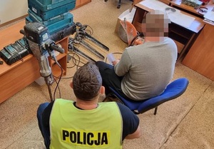 w policyjnym pokoju na krześle siedzi mężczyzna. Obok niego znajduje się nieumundurowany policjant w kamizelce odblaskowej z napisem Policja. Przed nimi na podłodze i na biurku leżą narzędzia elektryczne