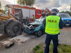 miejsce wypadku drogowego. Na chodniku stoi policjant ruchu drogowego. Przed nim znajduje się zielony samochód osobowy z wgniecioną maską oraz rozbitą szybą. Przed autem jest koparka. Łyżka koparki jest wbita w przód samochodu. Obok znajduje się wóz straży pożarnej oraz strażacy. Z tyłu stoi oznakowany policyjny radiowóz z włączonymi sygnałami błyskowymi