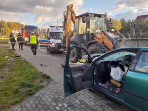 miejsce wypadku drogowego. Częściowo na chodniku i na jezdni stoi zielony samochód osobowy. Ma rozbitą szybę i wgniecioną karoserię z przodu. Poduszki powietrzne są wystrzelone. Wewnątrz auta chaos i ślady krwi. Przed autem stoi koparka, której łyżka jest wbita w przód samochodu. Policjant ruchu drogowego idzie w kierunku strażaków  i wozu strażackiego, przechodzi obok karetki