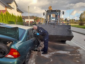 policjant służby patrolowej w rękawiczkach ochronnych nachyla się w kierunku wnętrza zielonego samochodu osobowego z wgniecionym przodem. Przed autem znajduje się koparka, której łyżka styka się z samochodem