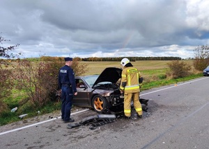 częściowo w rowie i na jezdni znajduje się ciemny samochód osobowy z wgniecionym przodem. Stoją przy nim policjant służby patrolowej oraz strażak. Na jezdni są porozrzucane fragmenty karoserii