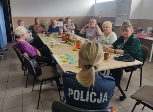 przy stole siedzą seniorki. Naprzeciw nich siedzi umundurowana policjantka