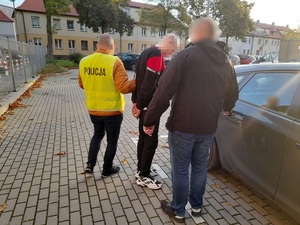 przed drzwiami samochodu stoi mężczyzna z rękoma skutymi kajdankami. Obok niego znajdują się dwaj nieumundurowani policjanci. Jeden z nich trzyma go za ramię