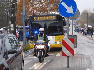 przed autobusem MZK jadącym na cmentarz jedzie policjant na motocyklu. Po obu stronach drogi poruszają się piesi