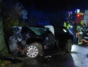 miejsce wypadku drogowego. Pora wieczorowa. Przy drzewie znajduje się rozbite auto z wystrzeloną poduszką powietrzną. Zderzak samochodu leży na jezdni. Przy aucie znajdują się dwaj policjanci. Jeden wypełnia dokumentację. Obok przy wozie straży pożarnej stoją strażacy