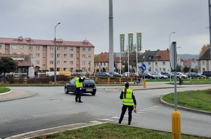 na rondzie stoi policjant ruchu drogowego, który pokazuje ręką sygnał do zatrzymania się Przy skrzyżowaniu stoi też policjantka. Skrzyżowaniem przejeżdża samochód osobowy. Przez przejście dla pieszych przechodzi kobieta.