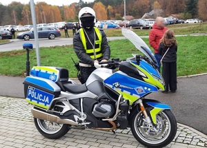 przy parkingu zapełnionym pojazdami i przed policyjnym motocyklem stoi funkcjonariusz ruchu drogowego. Za nim stoi mężczyzna z dziewczynką