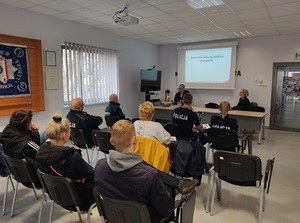 w auli budynku komendy policji przed ekranem na którym wyświetla się napis &quot;ochrona zwierząt podczas transportu&quot;, przy stole siedzą kobieta i mężczyzna. Przed nimi siedzą policjanci