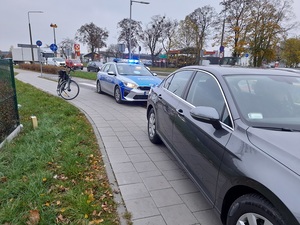 częściowo na jezdni i na chodniku zaparkowany jest grafitowy vw passat. Za nim tuż przy przejściu dla pieszych stoi policyjny radiowóz z włączonymi sygnałami błyskowymi. Obok radiowozu stoi rower