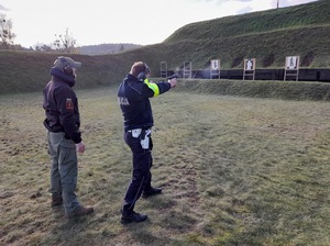 na wojskowej strzelnicy policjant ruchu drogowego oddaje strzał z pistoletu w kierunku tarczy. Za nim stoi instruktor