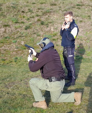 na strzelnicy wojskowej instruktor w pozycji klęczącej pokazuje stojącemu obok niego policjantowi przygotowanie krótkiej broni palnej do strzału