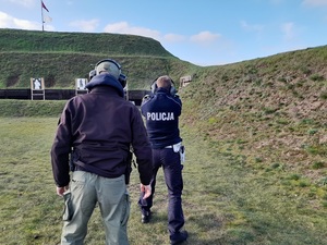 na wojskowej strzelnicy policjant ruchu drogowego oddaje strzał z pistoletu w kierunku tarczy. Za nim stoi instruktor