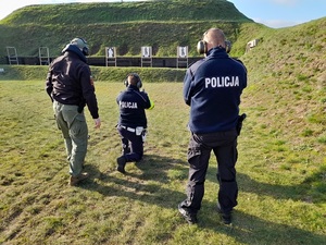 na wojskowej strzelnicy policjant w pozycji klęczącej przygotowuje się do oddania strzału w kierunku tarczy. Za nim stoją dwaj instruktorzy